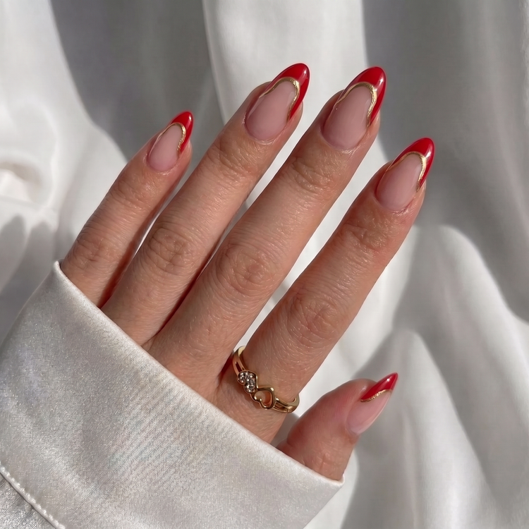 A close-up of a hand with long almond-shaped nails styled in a red French tip and nude pink base, wearing a delicate gold ring against a soft white fabric background.